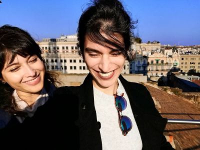 Sujaya Dasgupta is smiling with her friend as there are buildings in the background.